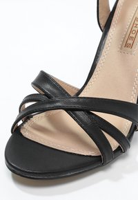 Sandale à talons en cuir noir avec des sangles croisées, un bout fermé et une semelle intérieure beige lisse avec des détails de couture subtils.