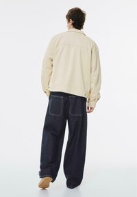 Beige oversized jakke med rygpaneldetalje, parret med mørkeblå vide jeans og beige sko, på en neutral baggrund.