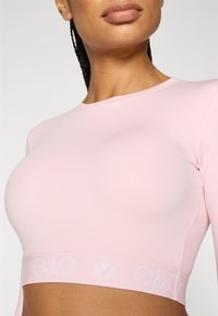 Ροζ μακρυμάνικο κοντό τοπ από λείο ύφασμα, με απαλό ελαστικό λουρί και λεπτομέρεια λογότυπου κατά μήκος του τελειώματος. Κοντινή προβολή του λαιμού.