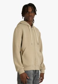 Felpa beige con zip, cappuccio con coulisse, tasche a marsupio e polsini a coste. Tessuto morbido con texture liscia e vestibilità casual.