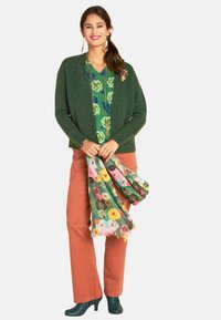 Grüne Strickjacke über einer blumigen grünen Bluse, kombiniert mit orangen Hosen. Hält einen mehrfarbigen Schal mit blumigen Mustern. Schwarze High Heels.