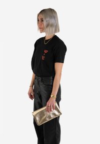 Musta t-paita, jossa on punaisia graafisia yksityiskohtia, yhdistetty korkeavyötäröisiin mustiin nahkahousuihin ja metallic-kultainen clutch-kassi kädessä. Lyhyt, suora hiustyyli.