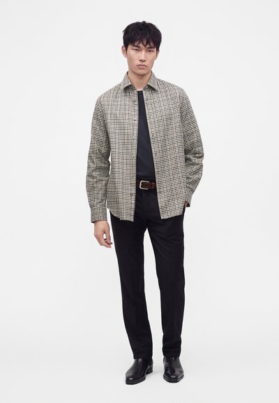 Νέος άνδρας με μαύρο πουκάμισο, καρό overshirt, μαύρο παντελόνι, καφέ ζώνη και μαύρα παπούτσια στέκεται σε λευκό φόντο.