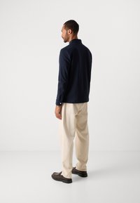 Man standing facing left wearing a navy long-sleeve shirt, beige trousers, and dark brown loafers on a plain white background.