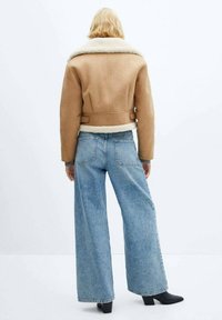 Tan croppet jakke med krave af shearling, kombineret med blå vidde jeans. Looket inkluderer sorte ankelstøvler og en minimalistisk baggrund.