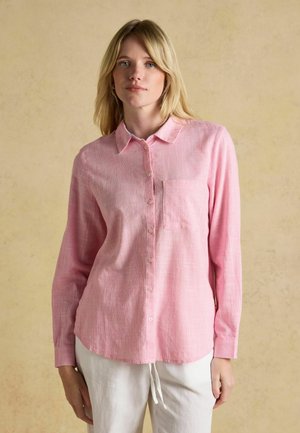 Žena oblečená v svetloružovej košeli s dlhým rukávom s náprsným vreckom a bielych nohaviciach, stojaca pred béžovým pozadím.