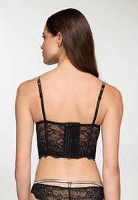Bralette in pizzo nero con spalline regolabili, motivo floreale, chiusura a gancio sul retro e bordi ondulati. Materiale morbido con una delicata texture.