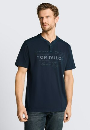 Mężczyzna z kręconymi włosami, ubrany w granatową koszulkę henley z krótkim rękawem z napisem "Tom Tailor" i współrzędnymi, stoi na tle jednolitego jasnego tła.