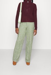 Persona que lleva pantalones de cuadros en verde y blanco, un suéter de punto burdeos, zapatos marrones y lleva un bolso de hombro verde claro.