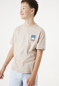 Beige Baumwoll-T-Shirt mit kurzen Ärmeln, das ein blaues rechteckiges Patch mit einem geschwungenen rot-weißen Design zeigt. Einfacher Rundhalsausschnitt, lässige Passform.