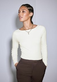 Light beige long-sleeve top made of ribbed fabric, paired with dark brown tailored pants. Gold chain necklace with layered design and accents.
