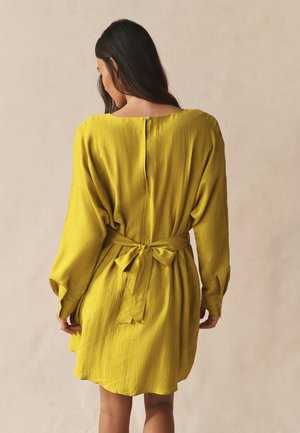 Kvinde med langt mørkt hår iført en løs sennepsgul kjole bundet med en sløjfe bagpå, står mod en ensfarvet beige baggrund.