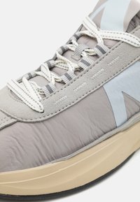 Grå treningsko med blandede materialer, inkludert semsket skinn og glatt stoff, med en beige såle og lyseblå detaljer.