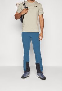 Muž v béžovém tričku North Face, modrých turistických kalhotách s černými záplatami, modrých turistických botách a nese černý batoh přes jedno rameno.