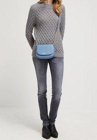 Maglione grigio a maglia a trecce, jeans slim grigi, stivali neri con tacco. Borsa a tracolla in pelle blu con catena dorata e dettagli del logo.