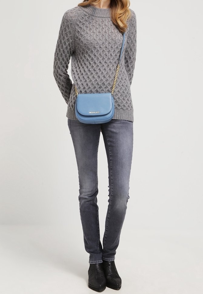 Maglione grigio a maglia a trecce, jeans slim grigi, stivali neri con tacco. Borsa a tracolla in pelle blu con catena dorata e dettagli del logo.