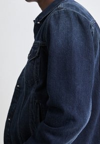 Chaqueta vaquera en azul oscuro, con cuello clásico, cierre frontal con botones, bolsillos laterales y detalles de costuras visibles en los hombros.