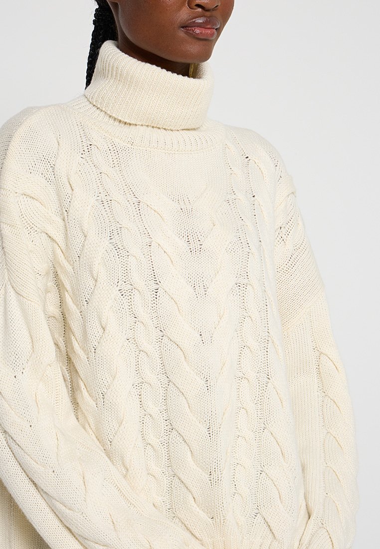 Grobstrickpullover in Creme mit hochgeschnittenem Stehkragen, Kabelstrickdesign, lockerer Passform und strukturierter Oberfläche, die einen gemütlichen Eindruck vermittelt.