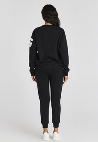 Kobieta stoi plecami, ubrana w czarną bluzę z numerem 6 na rękawie, dopasowane czarne dresy i białe trampki.