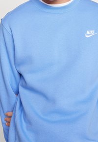 Sudadera azul claro hecha de tela suave. Presenta un cuello redondo y un logo blanco de Nike en la parte superior izquierda del pecho. Textura suave y diseño casual.