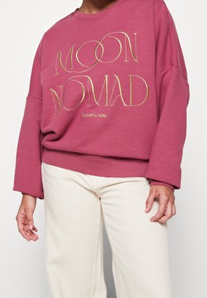 Personne portant un sweat-shirt rose ample avec "MOON NOMAD" en lettres dorées et un pantalon crème, debout devant un fond uni.
