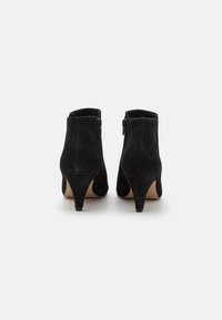 Ett par svarta mockaskor med anklar, med medelhöga avsmalnande klackar och sidoglittrar, visade bakifrån på en vit bakgrund.