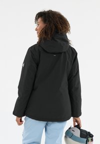Schwarze wasserdichte Jacke mit verstellbarem Kapuzen, lockerer Passform und Logo-Detail auf dem Ärmel. Die Person hält einen Helm in hellblauen Hosen.