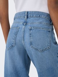 Pogled od zadaj osebe, ki nosi svetlo modre kavbojke z visokim pasom in velikimi zadnjimi žepi ter belo vloženo majico.