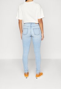 Mujer de pie con la espalda hacia la cámara, vistiendo una camiseta blanca, vaqueros ajustados de color azul claro y zapatillas naranjas sobre un fondo liso.