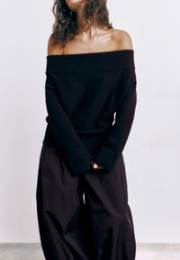 Vrouw met een zwarte off-shoulder trui en losse zwarte broek, staand tegen een effen witte achtergrond met de handen in elkaar gevouwen.