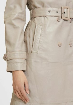 Trench coat in pelle beige con cintura in vita, bottoni doppiopetto e cuciture dettagliate su polsini e tasche.