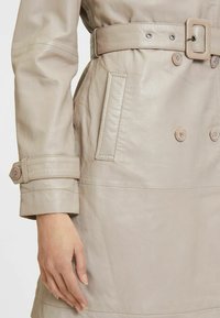 Trench coat in pelle beige chiaro con vita regolabile, bottoni doppi, e cuciture dettagliate su polsini e tasche.