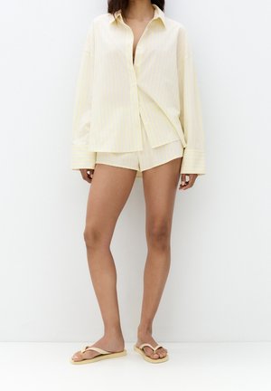 Femme portant une chemise ample jaune pâle à rayures boutonnée, un short assorti et des tongs beiges, debout sur fond blanc.