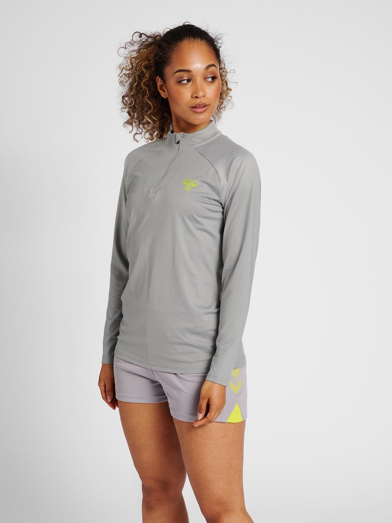 Femme aux cheveux bouclés portant un haut gris zippé à manches longues et un short gris assorti avec des accents jaunes, debout, regardant vers sa gauche.