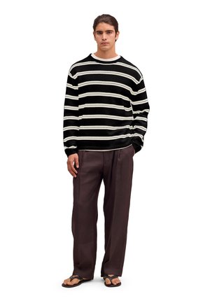Mand iført sort- og hvide stribede trøje, løstsiddende mørkebrune bukser og sorte flip-flops, stående mod en hvid baggrund.