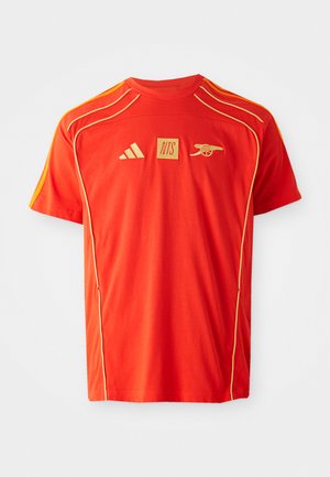 Rød kortermet T-skjorte med gule detaljer og piping. Har logoer: Adidas, NTS og Arsenal. Laget av mykt, pustende stoff.