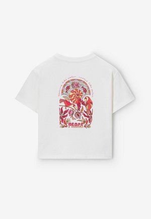 T-shirt blanc avec un design rétro coloré d'oiseaux, de plantes, de soleil et de poissons, comprenant les mots « Paix » et « Fort comme l'eau, enraciné comme un palmier ».