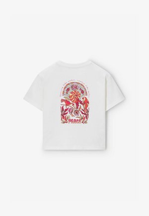 T-shirt blanc avec un design rétro coloré d'oiseaux, de plantes, de soleil et de poissons, comprenant les mots « Paix » et « Fort comme l'eau, enraciné comme un palmier ».