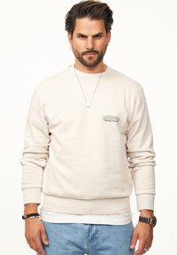 Beige Sweatshirt mit langen Ärmeln, gerippten Bündchen und Rundhalsausschnitt. Hat ein kleines Logo auf der Brust und ist über einem weißen Hemd getragen.