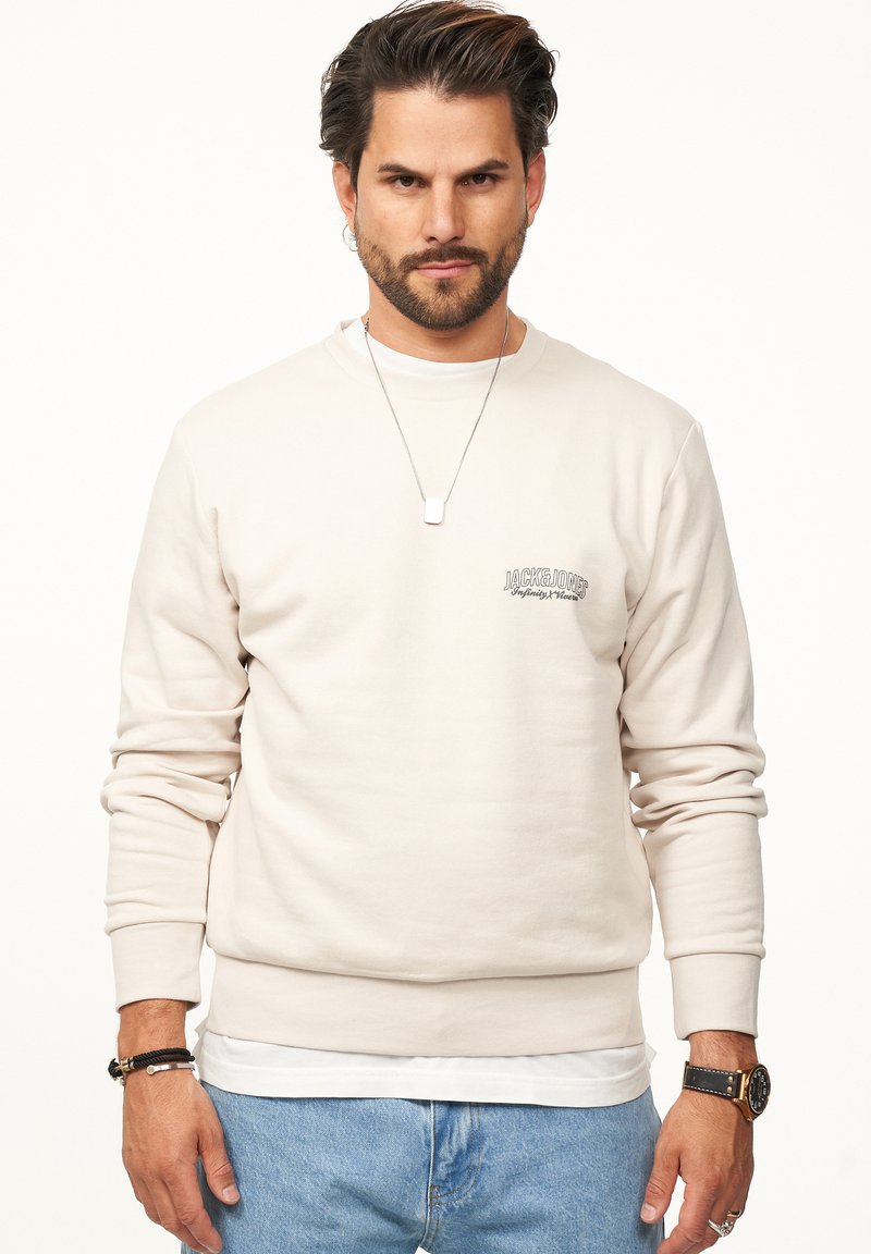 Beige Sweatshirt mit langen Ärmeln, gerippten Bündchen und Rundhalsausschnitt. Hat ein kleines Logo auf der Brust und ist über einem weißen Hemd getragen.