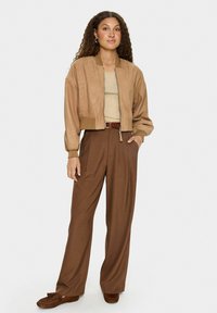 Frau mit lockigem Haar, die eine beige kurz geschnittene Jacke, ein hellbeiges Oberteil, braune weite Hosen und braune Mokassins trägt, steht mit einer Hand in der Tasche.