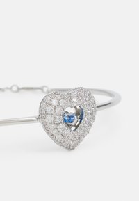 Bracciale a forma di cuore in argento con pietre trasparenti luccicanti, featuring una gemma blu al centro. Banda metallica semplice con dettagli della chiusura.