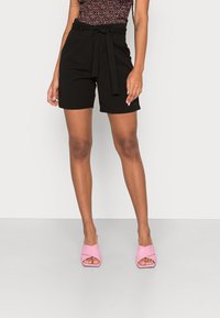 Pantalones cortos negros a medida con cinturón de lazo y bolsillos laterales, combinados con sandalias de tacón bloque abiertas en la parte delantera de color rosa. Tejido suave con un ajuste estructurado.