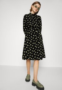 Abito nero con grandi pois verdi, dotato di colletto, chiusura a bottoni e una cintura in vita. Abbinato a mocassini verde chunky.