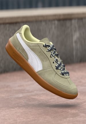 Beige ruskinds Puma-sneakers med hvid stribe, mønstrede snørebånd og gummisål, vist mod en sløret udendørs baggrund.