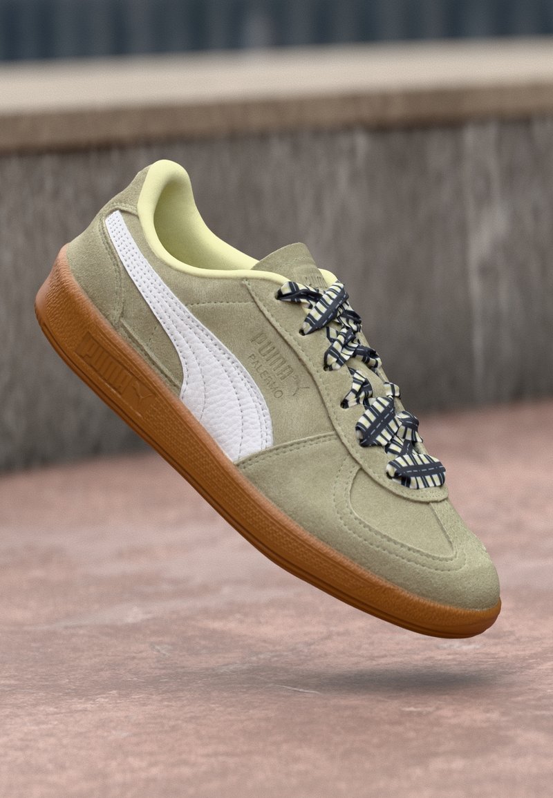 Baskets Puma en daim beige avec bande blanche, lacets à motif et semelle en gomme, présentées sur un fond extérieur flou.
