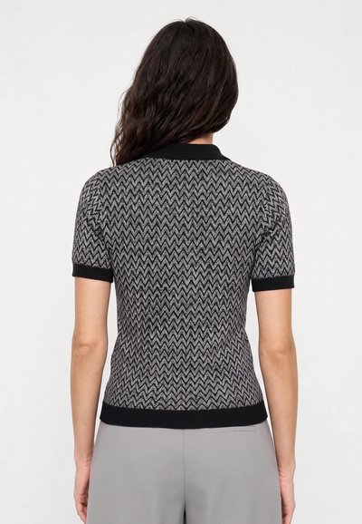 Femme aux longs cheveux foncés portant un haut noir et gris à manches courtes avec un motif en zigzag et un pantalon gris clair, vue de dos.