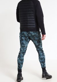 Černý svetr s prošívanými zády, doplněný o joggery s modro-zeleným maskáčovým vzorem, s žebrovanými manžetami a strukturovanými detaily. Černá obuv.
