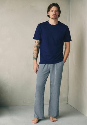 Mann steht barfuß an der Wand, trägt ein dunkelblaues T-Shirt und weiße Hosen mit dünnen schwarzen vertikalen Streifen, tätowierter Arm sichtbar.