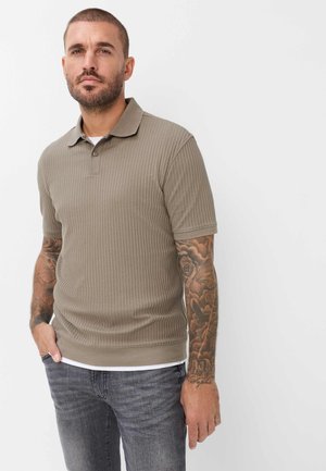 Man met kort haar en volle arm-tatoeages, gekleed in een beige kortemouw gebreide polo en donkere spijkerbroek, staand tegen een witte achtergrond.
